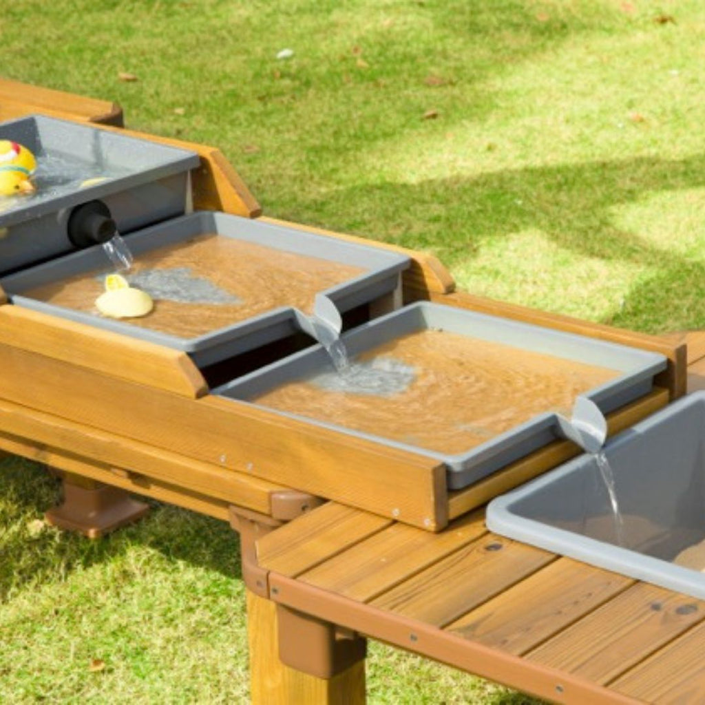 Kinderen spelen buiten met een houten water- en modderspeelset met meerdere speeltafels, watergoten en opvangbakken. Duurzaam buitenspeelgoed voor kinderdagverblijven en scholen, ideaal voor zand- en waterspel.
